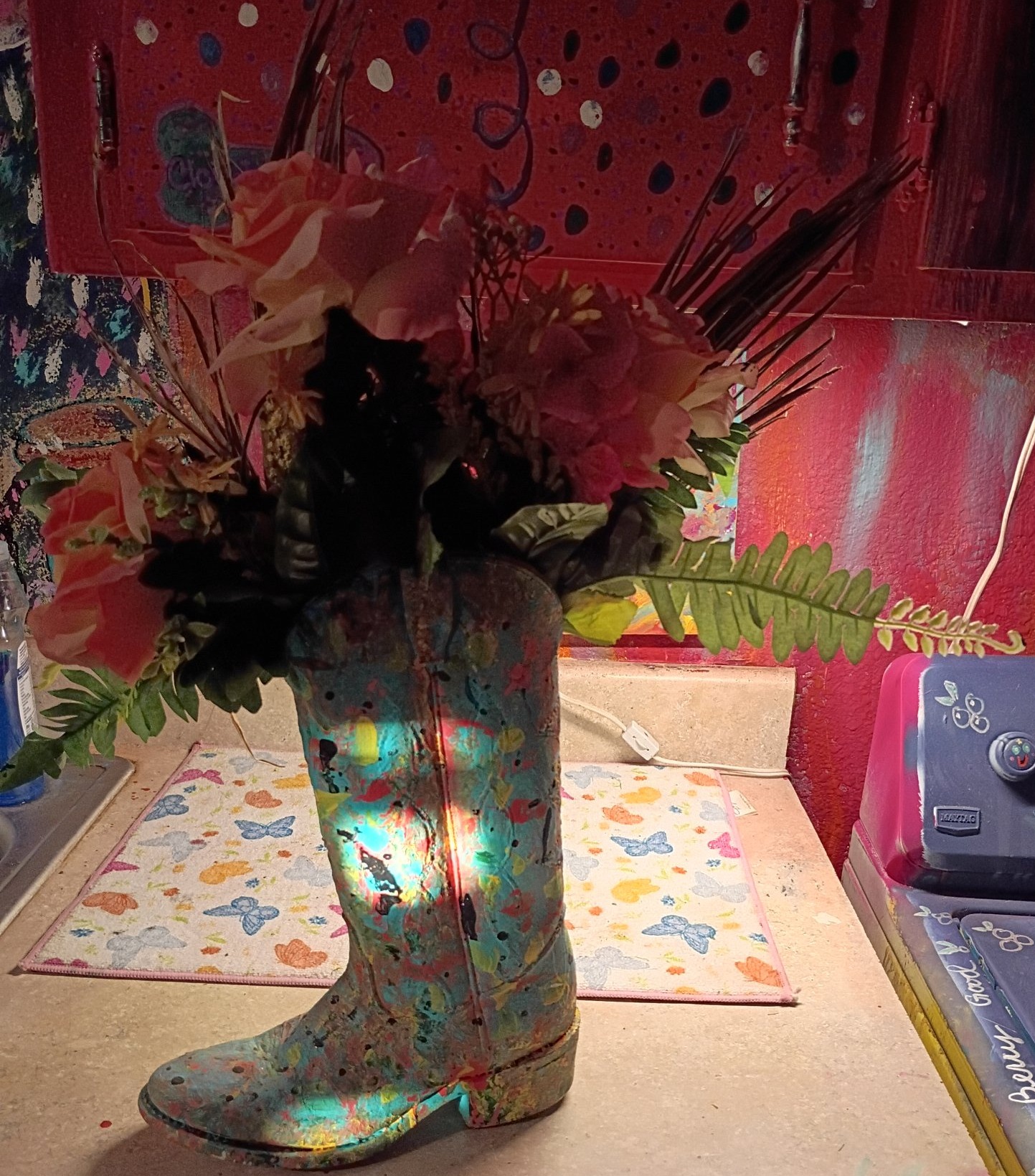 A hand-painted cowboy boot used as a vase — splashed in teal, pink and gold — holding pink roses and greenery