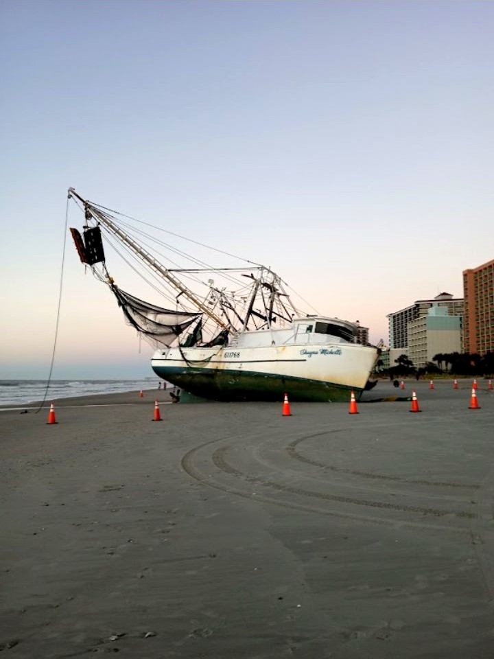 The Shayne Michelle still beached on the sand at dawn, ringed with orange traffic cones, the ocean to one side and beachfront hotels behind her