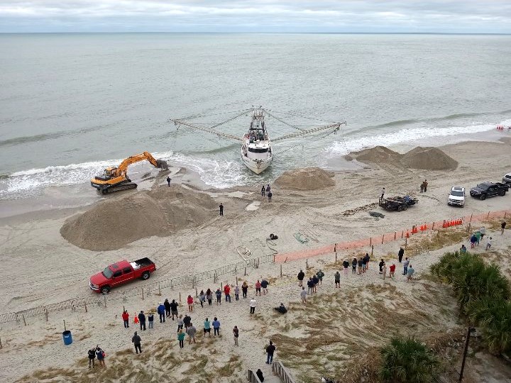 From above: the Shayne Michelle back at the water's edge while a yellow excavator digs her out, piles of sand around her and a crowd watching from behind a fence