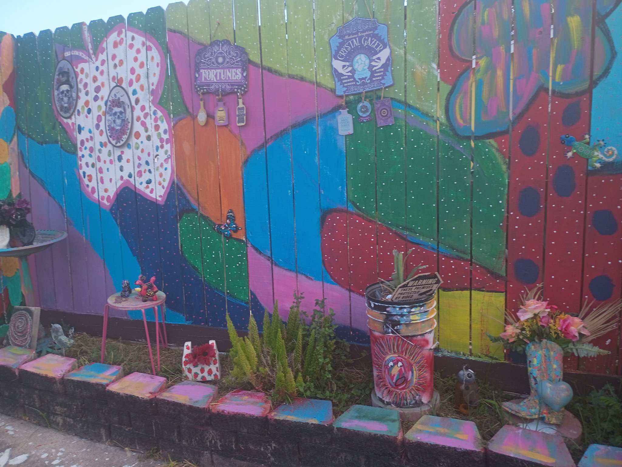 Mia Roze Bootz's fence mural with her painted garden pieces in front of it — a hand-painted bucket planter with a sun face and a painted cowboy boot holding flowers, along a border of painted blocks