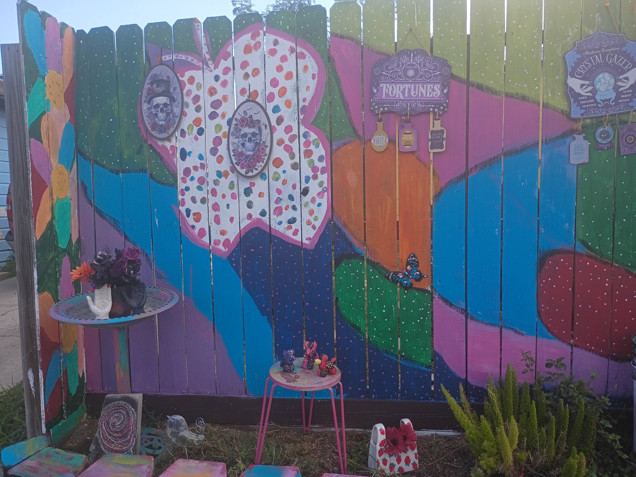 Mia Roze Bootz's fence mural, wide view — a giant white butterfly covered in multicoloured dots painted across a wooden fence in pink, teal, green, orange, and purple, with Fortunes and Crystal Gazer signs hanging on it