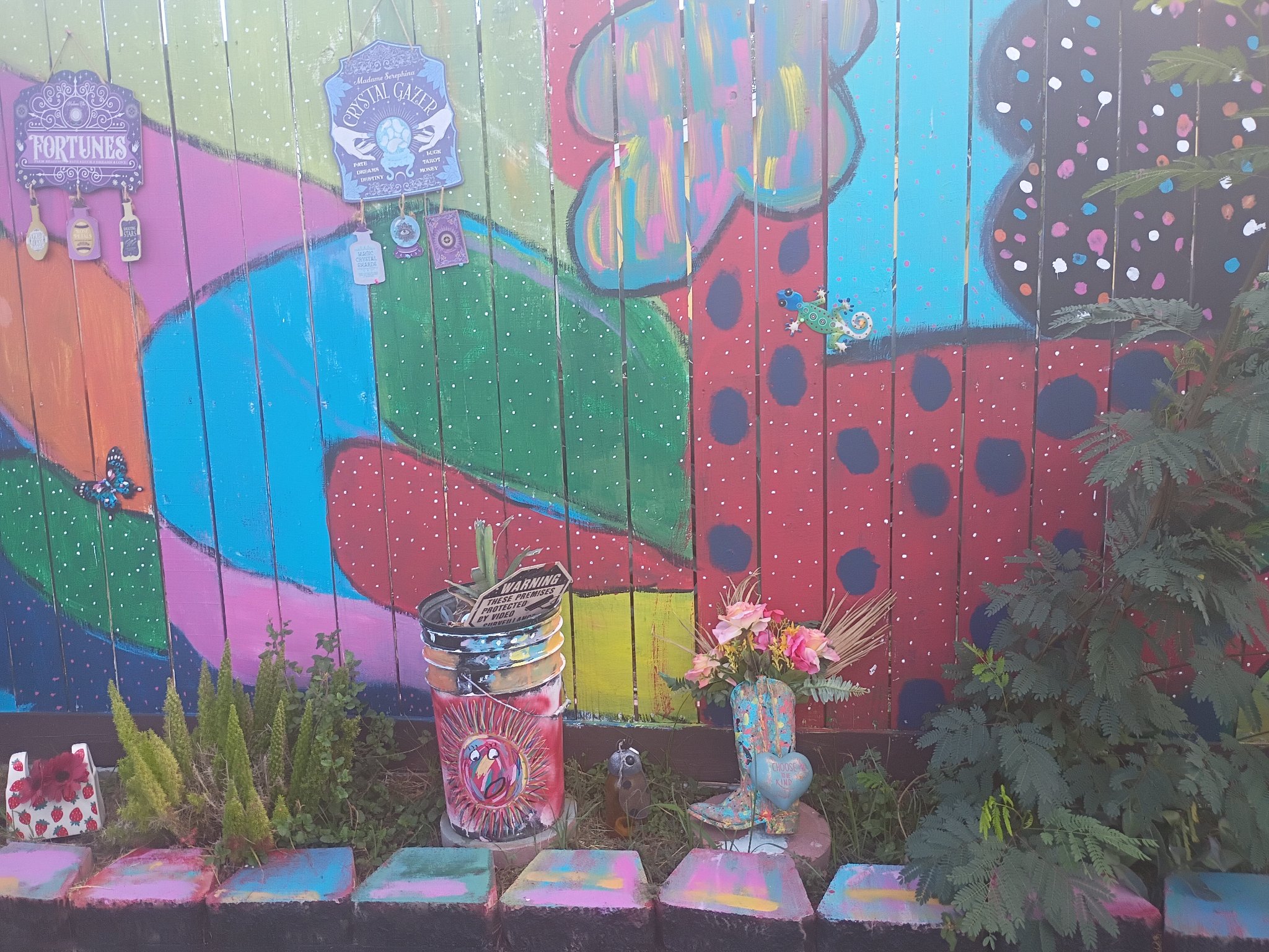 Right section of Mia Roze Bootz's fence mural — bold red with dark polka dots, teal and green waves, white star specks, and a small painted gecko, with the painted bucket and boot planter below