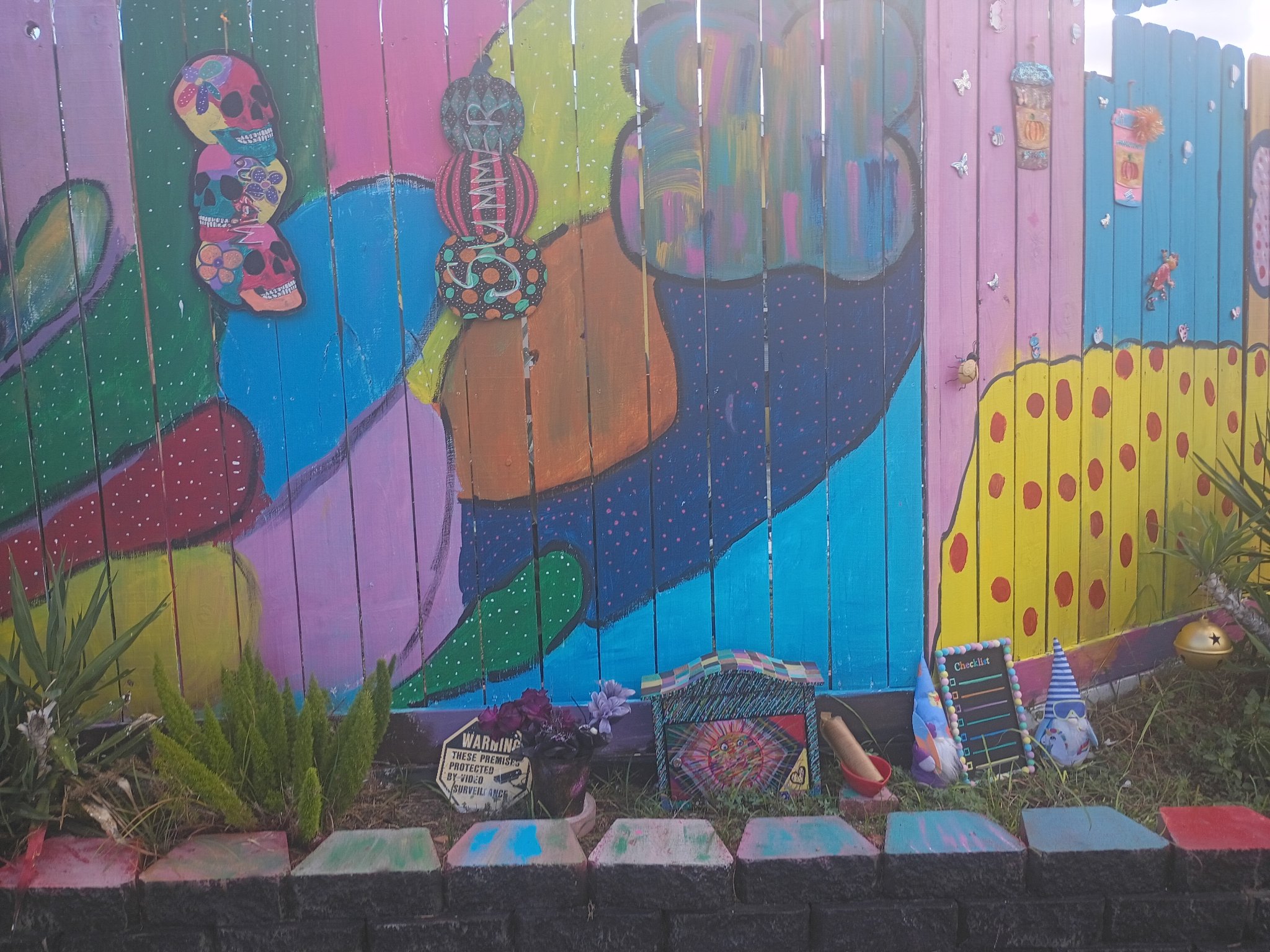 Mia Roze Bootz's fence mural with colourful stacked skulls and a Summer sign hanging on it — sweeping shapes in teal, pink, green, orange, and yellow beside a yellow fence panel with red polka dots