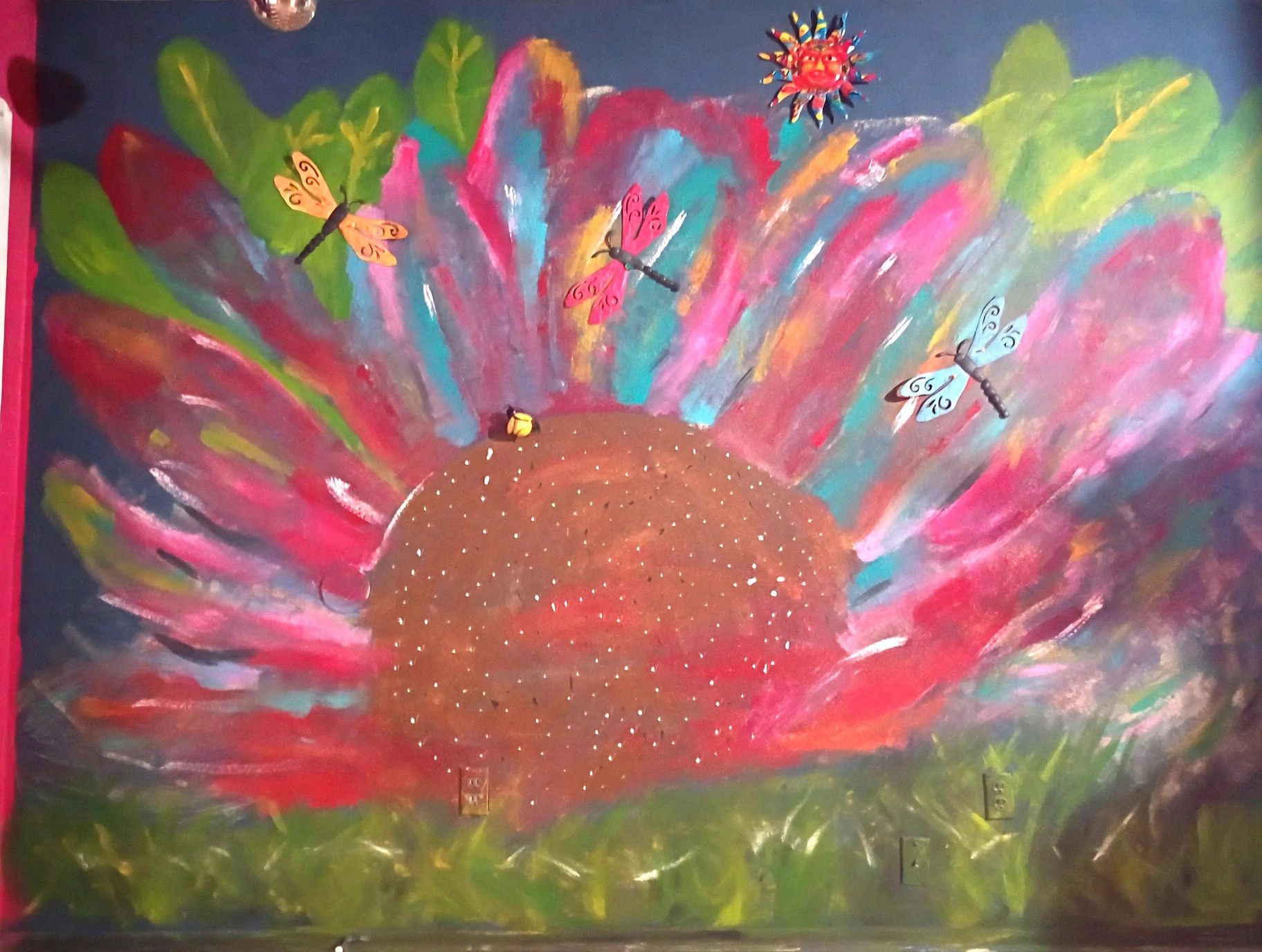 Mia Roze Bootz's sunset wall mural — a large glitter-dotted sun rising over green grass, its rays sweeping out in hot pink, red, teal, and orange, with three dragonflies in gold, pink, and blue flying across it