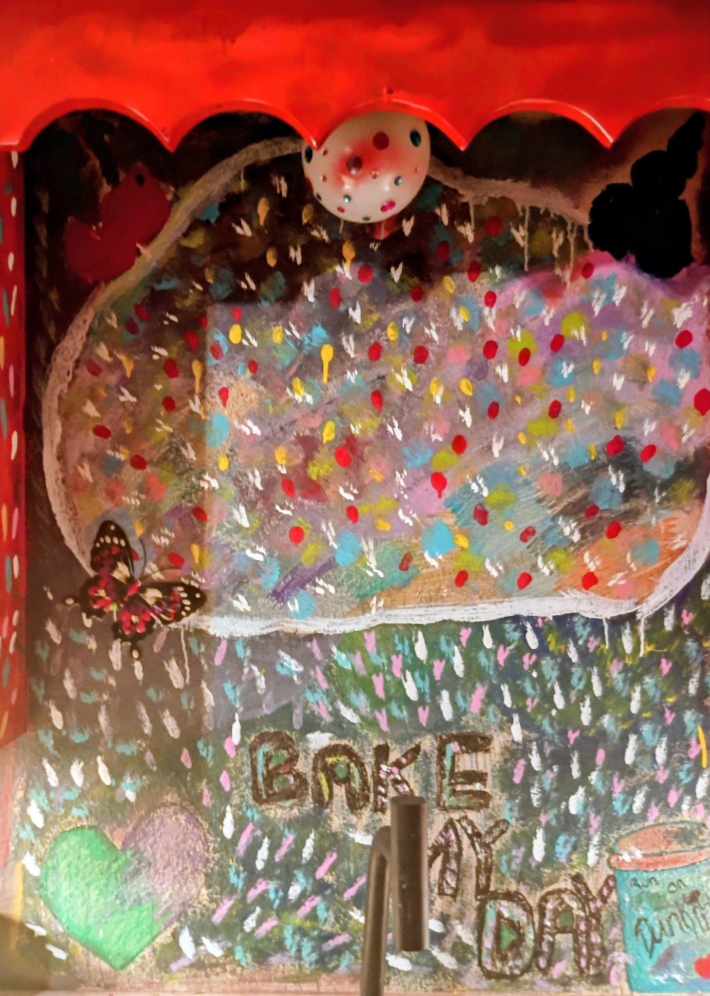 Inside of Mia Roze Bootz's painted kitchen cabinet — a cloud covered in colourful sprinkles with a butterfly at its edge, a painted heart, and the words Bake My Day, under a red scalloped cabinet top with a polka-dot knob