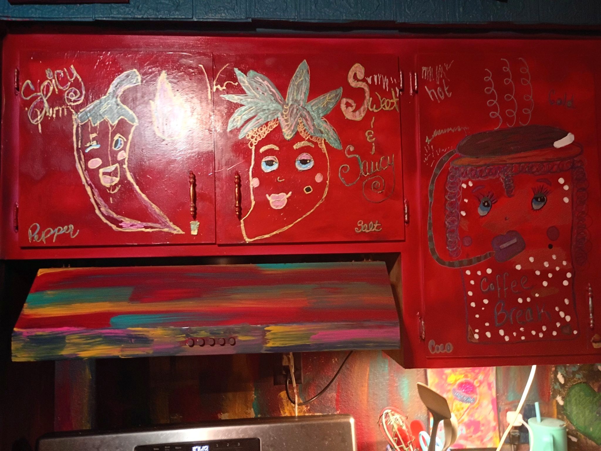 Mia Roze Bootz's painted kitchen cabinets — deep red cabinet doors hand-painted with a smiling chili pepper labelled Spicy Yum, a sweet pepper labelled Sweet and Saucy, and a coffee cup character labelled Coffee Break, above a range hood painted in rainbow stripes