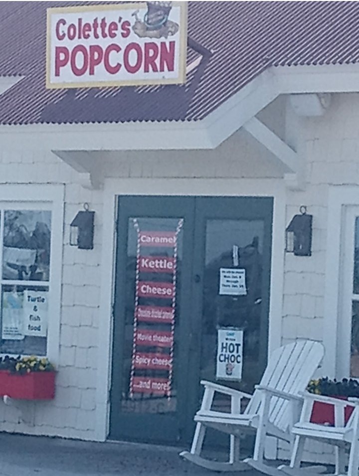Colette's Popcorn shopfront — a white shingled shop with the Colette's Popcorn sign over green double doors listing caramel, kettle, and cheese, with white rocking chairs on the porch