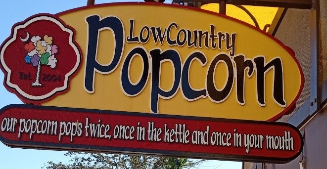 The LowCountry Popcorn sign — a yellow oval reading LowCountry Popcorn beside a red badge with a palmetto tree of popcorn, over a red banner reading our popcorn pops twice, once in the kettle and once in your mouth