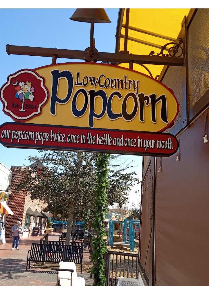 The LowCountry Popcorn hanging sign — a yellow oval with a palmetto tree of popcorn, over a red banner reading our popcorn pops twice, once in the kettle and once in your mouth